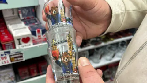 Kensington & Chelsea Council A man holds a stack of three glass shot glasses which have an artistic drawing of paddington bear waving his red hat, as well as Big Ben and Tower Bridge on them. Shelves of shop merchandise can be seen in the background