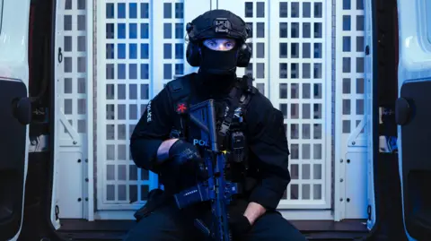 BBC/Expectation Entertainment/Drew Forsyth An armed police officer, holding his gun in a downward position and wearing a mask, looks toward the camera while sat in the back of a police van.