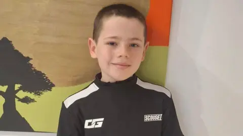 Family photo Phoenix has short dark hair and is wearing a sports top. He is standing in front of a canvas which is hanging on the wall.