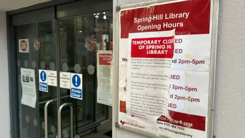 The opening hours are taped over by a closure notice. There are modern power assisted doors with a keep clear sign. 