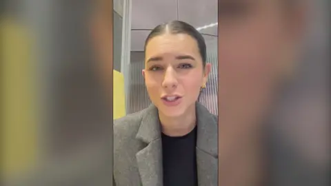 A woman with tied back brown hair wearing a grey blazer looks at the camera 