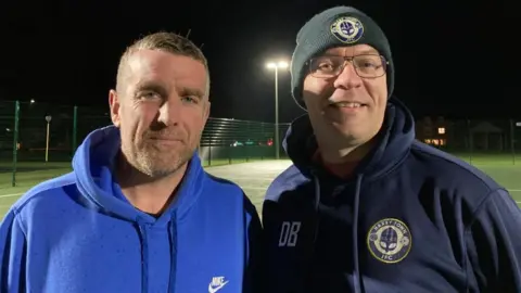 Matt Boggan and David Blackmore stand side by side, in front of sports courts. Matt has short brown hair and stubble, and wears a blue Nike branded hoodie. David wears a Haxby Town JFC branded woolly hat, glasses and a dark blue hoodie with the Haxby Town JFC logo on the chest.