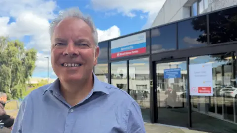 BBC/Jim Scott Dr Alan Sweenie, who is wearing an open blue-collared shirt, is at the entrance of the NHS Blood and Transplant centre in Newcastle. The entrance is clad with black glass and features signs urging people not to smoke, and that they are in the 'main site entrance'.