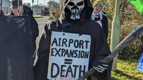 One protester dressed in a halloween costume with a sign on it that read "airport expansion = death"