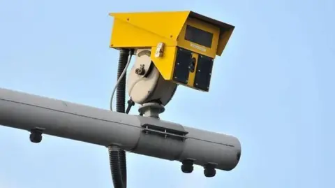 A yellow ANPR camera connected to a grey post with blue sky in the background