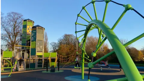 An image of a large green play area, with a large metal slide and netted bridge