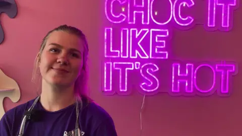 A young woman wearing an apron smiles at the camera. Behind her is a neon sign saying "choc it like it's hot" to advertise her baking business.