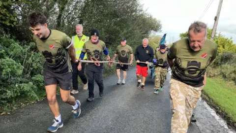 BBC People pulling a field gun up a hill. They are all holding rope and working together to pull it up the hill. Either side is grass.