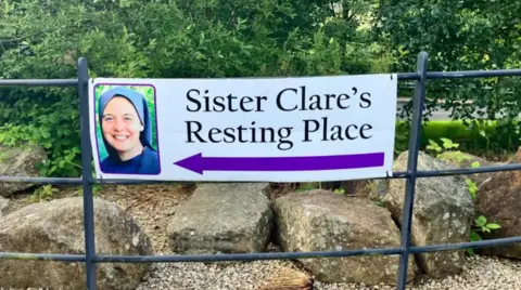 A sign reading: ' Sister Clare's Resting Place' and a purple arrow pointing left is attached to a black railing. A number of trees and stones can be seen in the background.