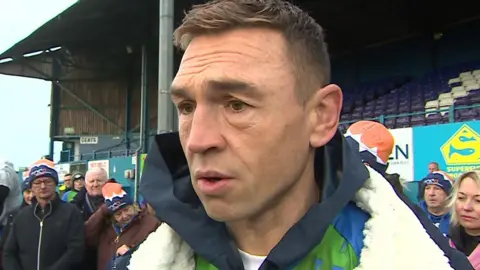 Kevin Sinfield at the Fibrus stadium in Workington, being interviewed by a reporter who is out of shot. He has short brown hair and green eyes. He is wearing a fleece-lined blue coat over a green and blue sports top. Behind him is a group of supporters.