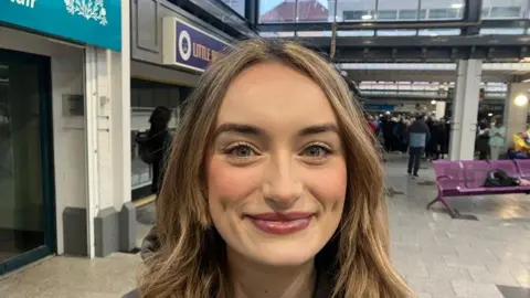 Bernadette Hagans, a woman with brown fair hair down to her shoulders. She is standing in the centre of a shopping centre, closed shops and a purple bench can be seen behind her.