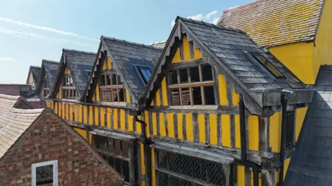 The restored building, it is yellow with black stripes, it has five adjacent gables with black tiled rooves.