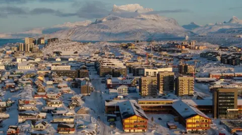 Reuters A drone view shows a view of houses and high rise buildings in the snow with a snowy mountain high above in the background, in the capital of Greenland, Nuuk, on 15 January 2026. 
