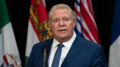 Doug Ford stands before a black microphone. He is wearing a navy suit, white shirt and blue tie. He is looking straight ahead. Behind him are several colourful flags.