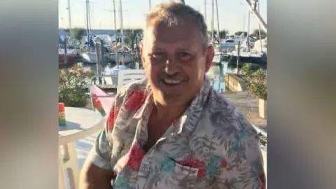 Family photo Mike O'Leary wears a tropical shirt, smiling in the sunshine at a harbour, boats visible in the background