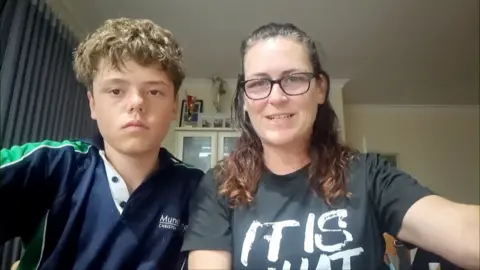 Austin Applebee, who has blonde curly hair and is wearing a navy top with green stripes down the arm, sits next to his mother Joanne, who wears glasses and has brown hair, in a living room