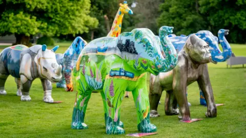 Sculptures of animals, including two elephants, two gorillas, a rhino, and a giraffe, are lined up on grass. They are all painted in different colours with designs on them.