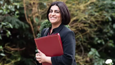  Home Secretary Shabana Mahmood walks outside of Downing Street. She wears a high-necked black jacket, black bobbed hair, and holds a burgundy folder.