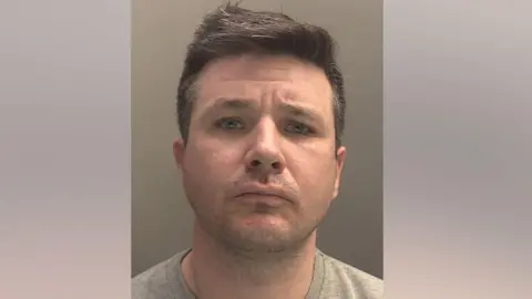 A police mugshot of Stephen Bates, 42, who has brown hair and is wearing a grey t-shirt, and is looking into the camera with a concerned expression.