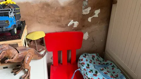 Jessica Pourasghar Children's toys sit infront of a wall that has had its paint removed and has mould stains