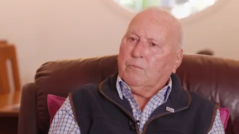 BBC An elderly man wearing a white shirt and blue fleece. He is sat in a brown leather chair looking to the left of the camera