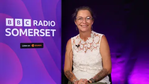 Dawn Carey, who is wearing a white lace top and has tattoos on her lower arms, is sitting down next to a purple BBC Radio Somerset banner.