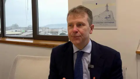 Matt Thomas, a man wearing a dark blue suit, light blue shirt and royal blue tie. He has short brown hair. He is sitting in front of a print of La Corbiere Lighthouse, and a window through which the marina is visible.