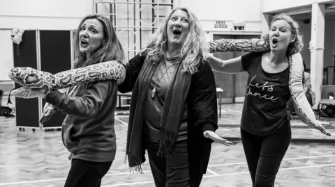 Cat Humphries Three women taking part in a rehearsal. Their mouths are open and their arms are out and they are holding a toy snake. The image is black and white and the rehearsal is taking place in a wall with sport equipment behind. 