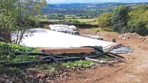 Bradford Council A hillside construction site with a partially built platform covered in white sheeting, scattered pipes and materials in the foreground, and a wide valley of fields and rolling hills stretching out under a partly cloudy sky.