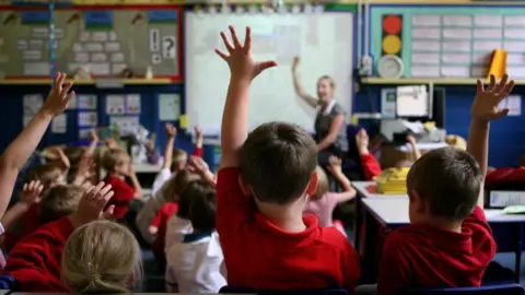 PA Media Young children in a classroom wearing red and white tops. Some have their hands up while a teacher is at the front of the room in front of a white board.