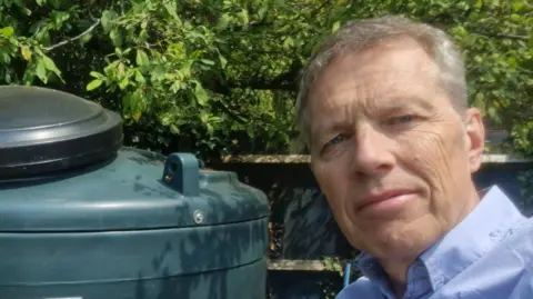 A man in a blue shirt takes a selfie in a garden next to a green oil tank. He has grey hair and there are leaves behind the tank.