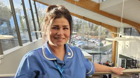 A woman with brown hair tied up in a bun, and brown eyes, looks into the camera, smiling. She is wearing a blue nurse uniform with a lanyard that has the NHS logo on it. She is stood inside a glass building with her arm resting on a metal railing behind her.