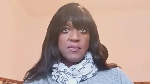 BBC Grace Gomez, a black woman with long dark hair, is pictured facing the camera. She is wearing a white scarf with images of the Christian cross on it. 