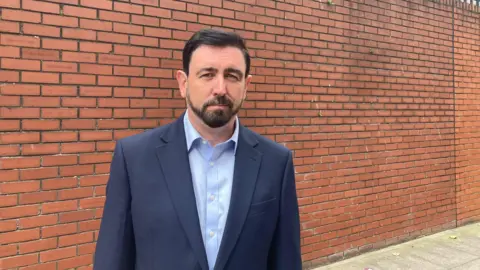 A man in a blue suit, light blue shirt is stood in front of a red brick wall. He has dark hair and a black beard. He is looking directly at the camera