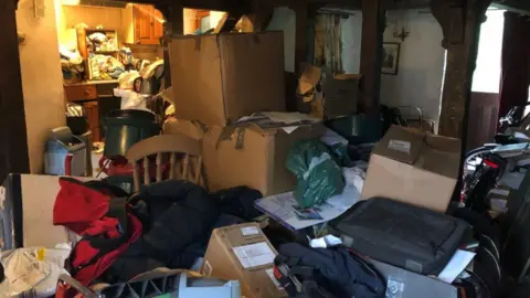 A kitchen and living room area filled with clutter. The floor cannot be seen and there are large boxes piled up among the clutter.