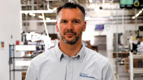 Oliver Conger smiles at the camera. The medium close-up is taken at his factory, and some workstations are out of focus in the background. He wears a pale blue shirt that features the dark blue logo of British Rototherm on his chest.