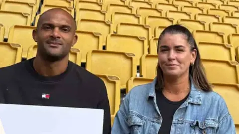 Wolves Foundation A man with a shaved head wearing a black sweater sits next to a woman wearing a light blue denim jacket in a football stadium. Behind them are rows of yellow seats