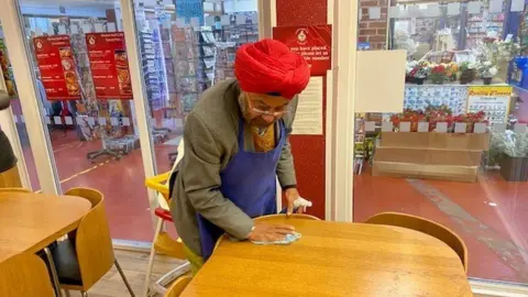 A man is wiping down a wooden table inside a café. He is wearing a blazer, blue apron and red turban.