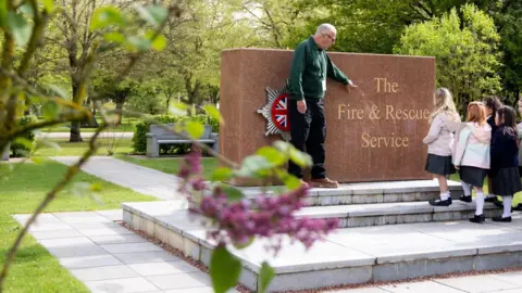Staffordshire County Council A man is standing on a stone step and is pointing at a memorial plaque that says 'The Fire and Rescue Service'. In the background, a path winds throughout trees.