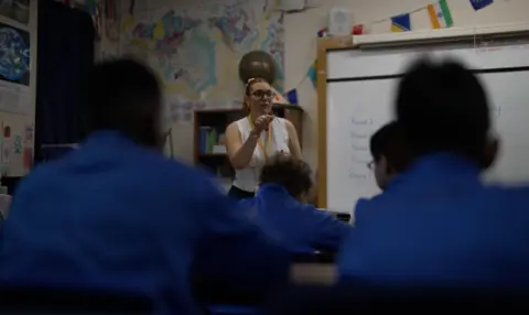 BBC/BRIJESH PATEL A private school teacher stands in front of a classroom looking down at the class who are sitting a tables in a row with their backs to the camera