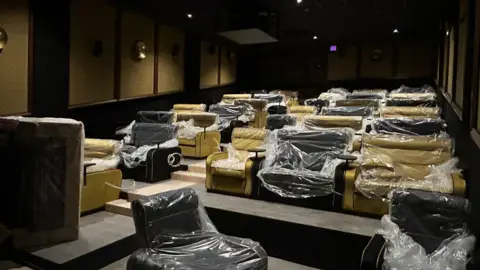 A small cinema with yellow and black plush armchairs, which are covered in plastic wrapping. The photo is taken from the end with the screen, looking up at the seats.