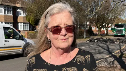 A woman with blonde hair wearing sunglasses with a road in the background where work to remove speed humps is taking place.