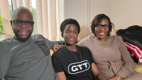 Sam Read/BBC Daniel Chenjerai, known by the stage name DC3, sits on a sofa between his mother Annastacia Chenjerai and father Pardon Chenjerai.