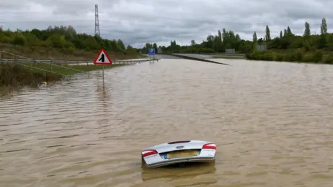 PA Media The open boot of a car is visible above the water where the vehicle is submerged in flood water on a421 in Marston Moretaine