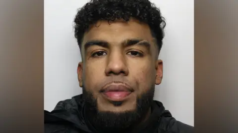 A police mugshot of a man with curly black hair and a neat black beard. He is wearing a black coat