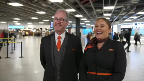 Ant Saddington/BBC Two smartly dressed people in black, with flashes of orange, are smiling and laughing in an airport terminal.