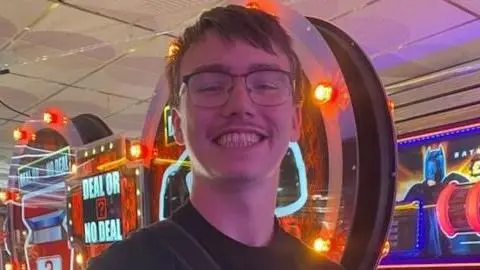 Luke Cusack was pictured grinning at the camera while standing in an amusement arcade. He had short brown hair and wore glasses and a black T-shirt.