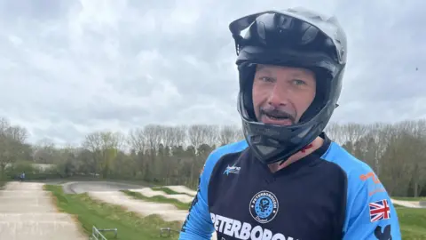 Connor Bennett/BBC Tim Stratford, a rider in a light and dark blue top saying Peterborough Phantoms. She is wearing a helmet and starting to smile. On his right is the undulating concrete track. Behind it are leafless trees under a grey sky.