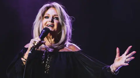 Bonnie Tyler on stage singing. She wears a black long sleeve dress and holds the microphone in one hand, with the other outstretched. Her long blonde hair is loose. 