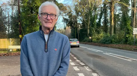 Clare Worden/BBC Brian Shaw stands with the busy A148 road behind him. He has white hair and wears black glasses and a blue top. 